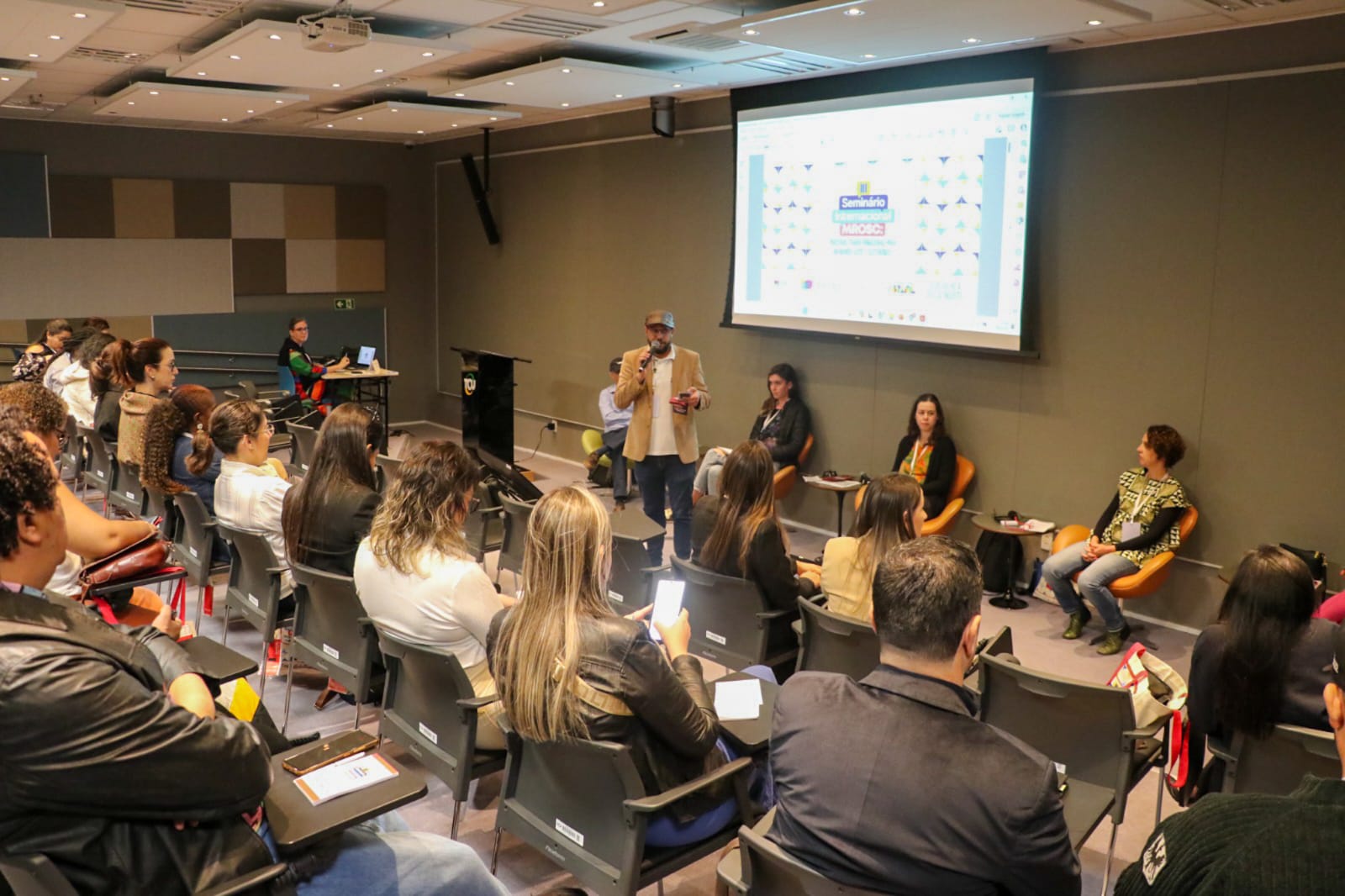 Na foto vê-se uma planteia em um auditório e um homem ao centro falando.